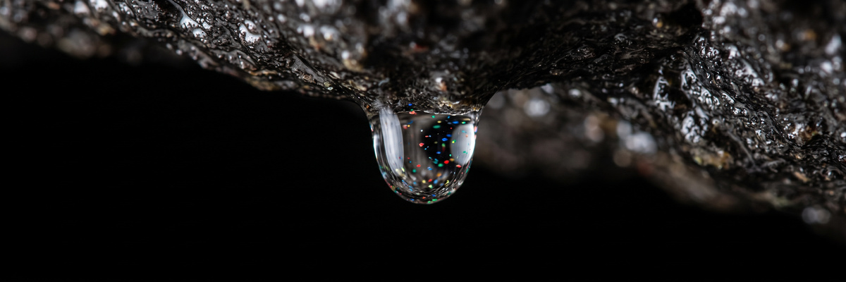 Water droplet hanging from dark aquifer rock with tiny multicolored particles suspended inside